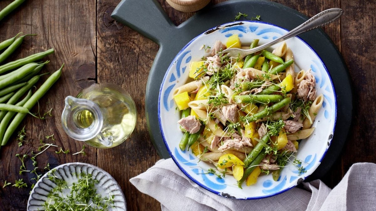 Ein weißer Teller mit blauen Verzierungen gefüllt mit einem Nudelsalat mit Bohnen und Thunfisch auf einem hölzernen Hintergrund. Darunter ein hellgraues Geschirrtuch. Links davon eine kleine Karaffe mit Öl, eine kleine Schüssel mit Kresse und grüne Bohnen.