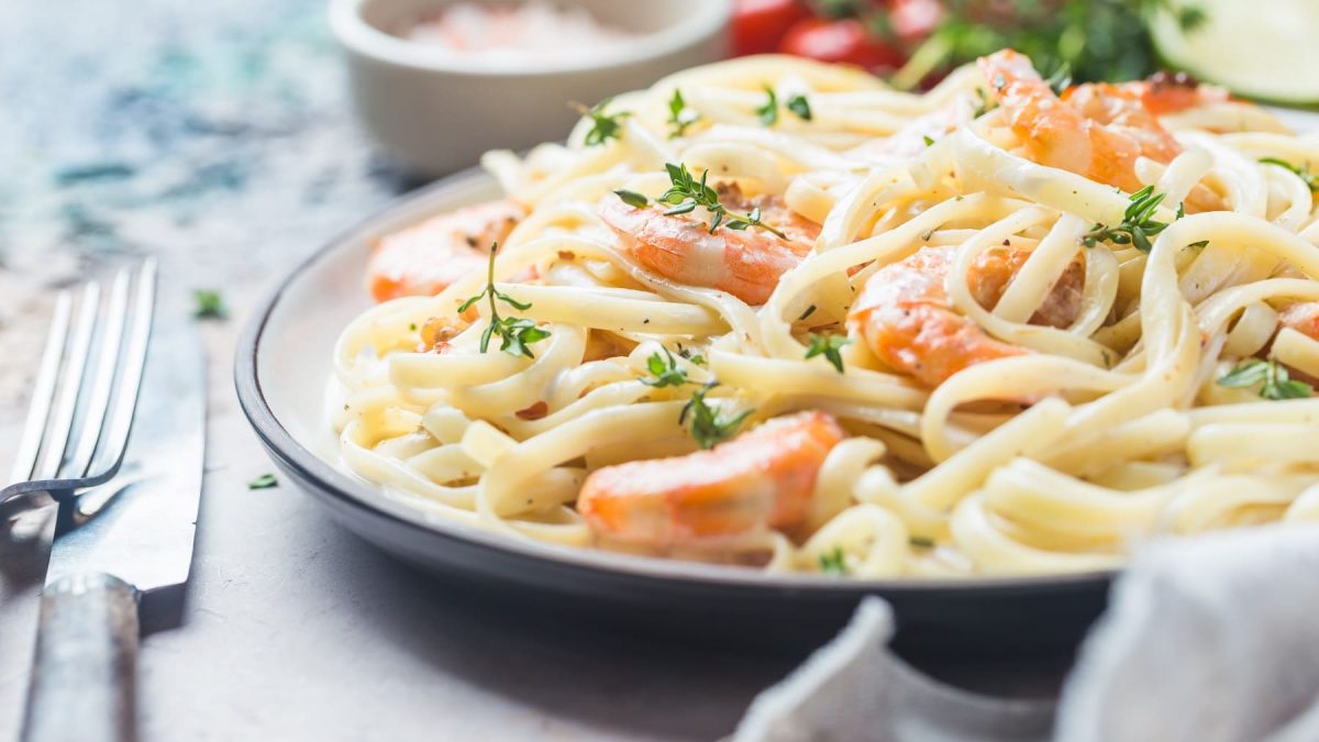 Ein weißer Teller mit blauem Rand und der Pasta mit Lachs und Garnelen auf einem grauen Untergrund sehr nahe fotografiert.