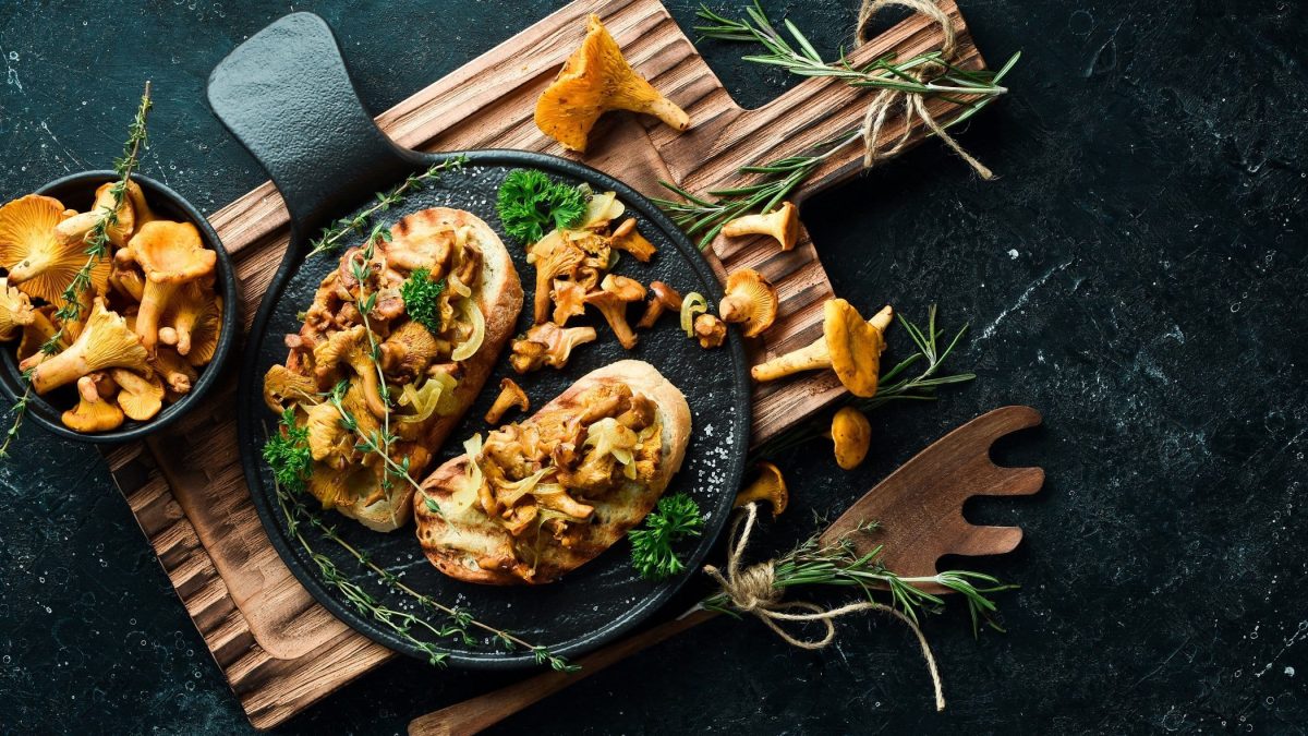 Draufsicht: Dunkler Untergrund, zwei Pecorino-Bruschetta mit Pfifferlingen auf einem Holzbrett und drum herum frische Pfifferlinge und frische Kräuter.