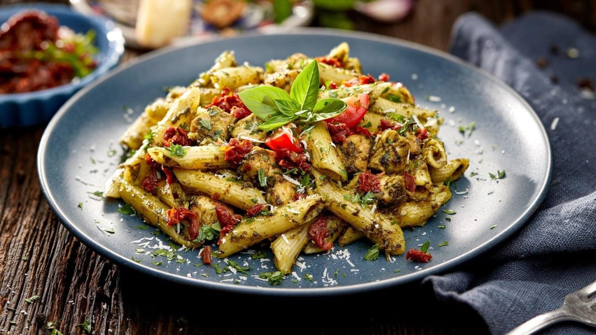 Draufsicht, schräg: ein blauer Teller Pesto-Pasta mit getrockneten Tomaten steht auf einem Pesto-Pasta mit getrockneten Tomaten. rechts neben liegt Besteck.