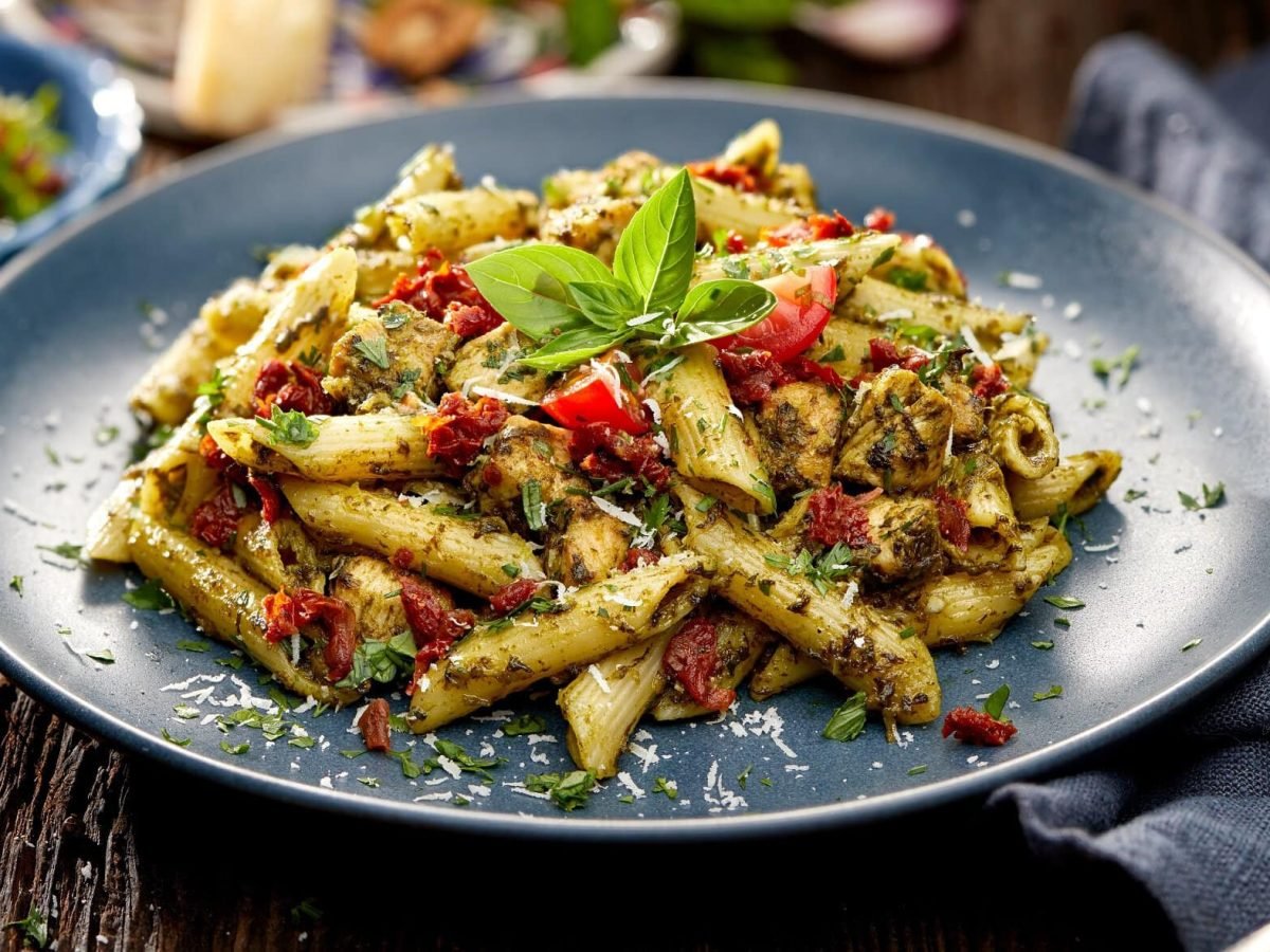 Draufsicht, schräg: ein blauer Teller Pesto-Pasta mit getrockneten Tomaten steht auf einem Pesto-Pasta mit getrockneten Tomaten. rechts neben liegt Besteck.