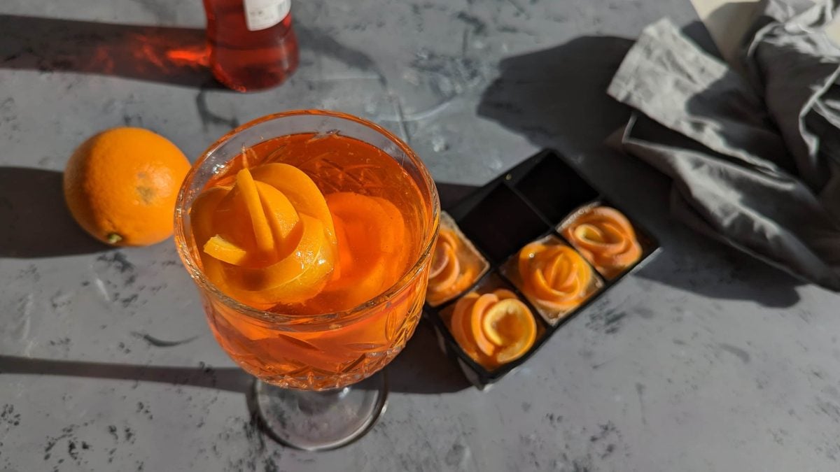 Ein Glas Aperol Spritz auf grauem Untergrund mit einer Aperolflasche und einem Aperol Spritz.