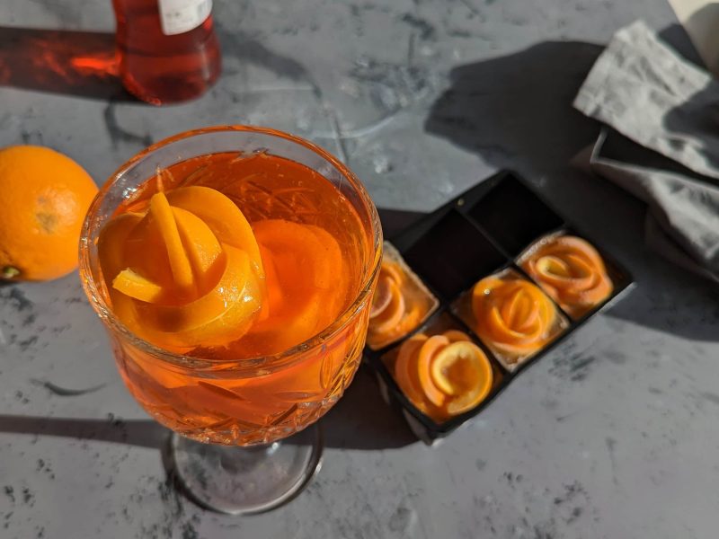 Ein Glas Aperol Spritz auf grauem Untergrund mit einer Aperolflasche und einem Aperol Spritz.