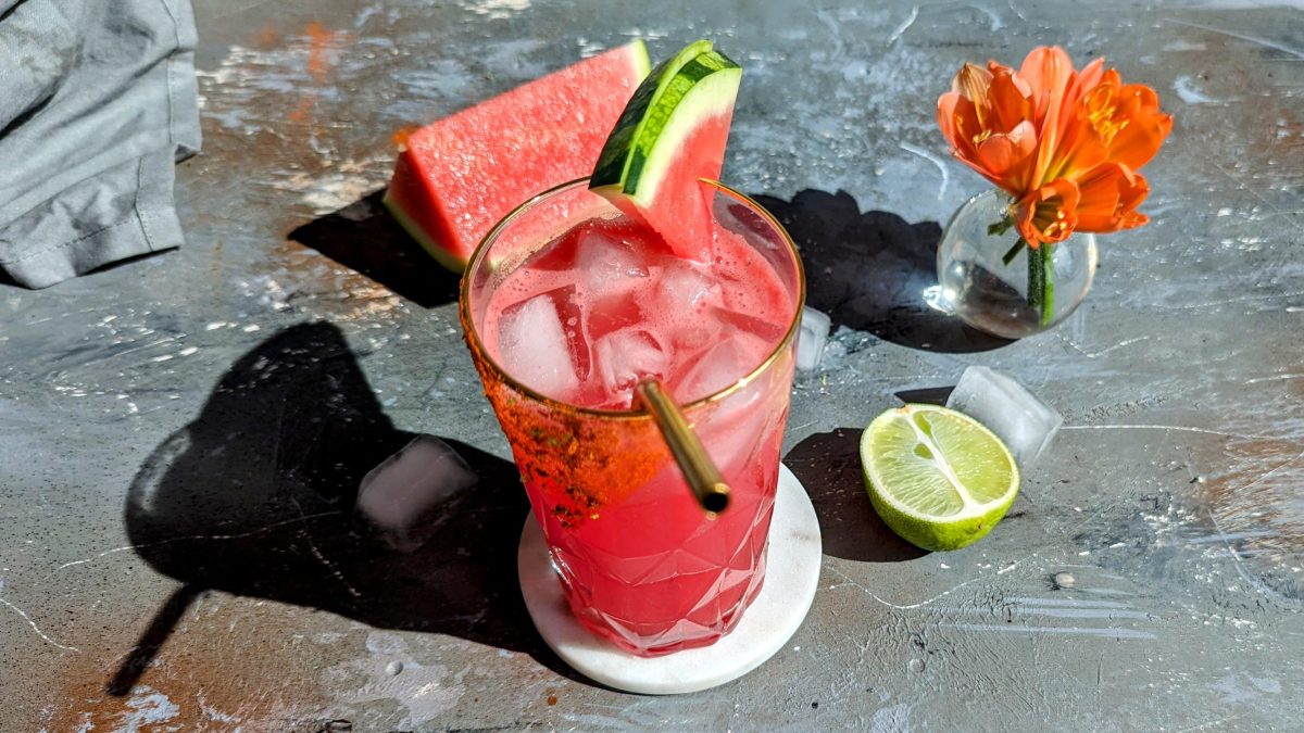 Ein glas mit dem pinken Watermelon-Paloma auf einem grauen Tisch mit rötliche rkleiner Pflanze, Eiswürfeln, halber Limette und grauer Serviette.