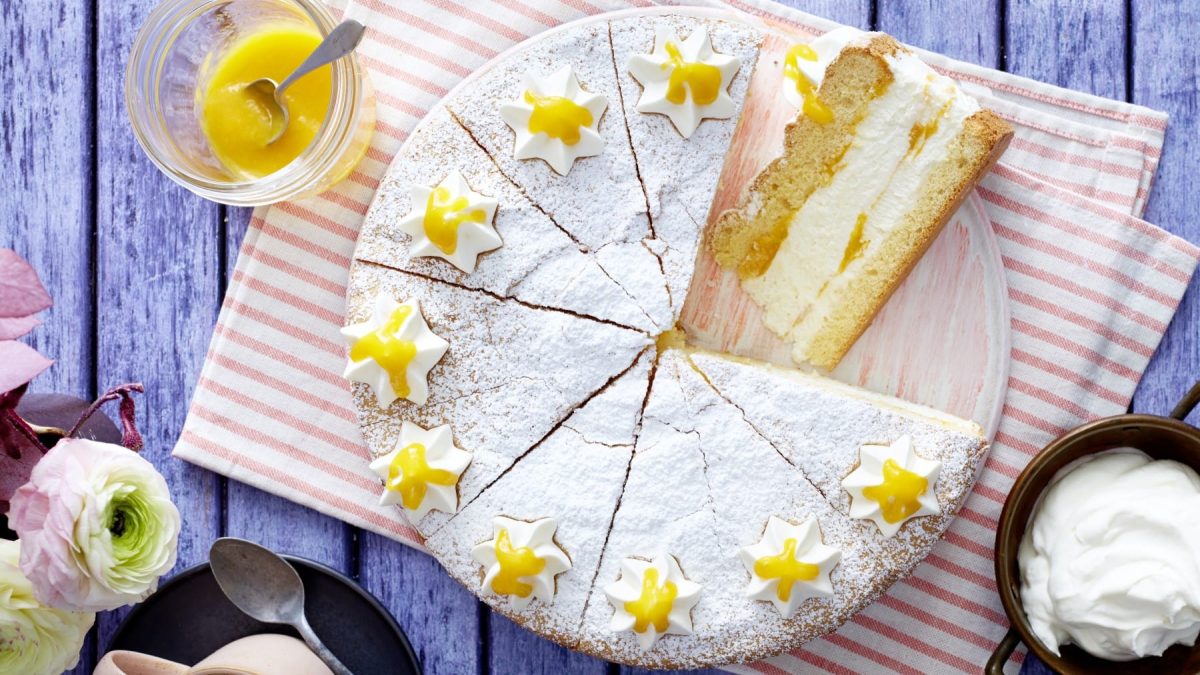 Draufsicht: Angeschnittene Quark-Mangotorte auf einem Teller, darunter ein rot-weiß-gestreiftes Küchentuch auf blauem Holzuntergrund, daneben in Schälchen Mangopüree und Sahne, sowie Blumen.