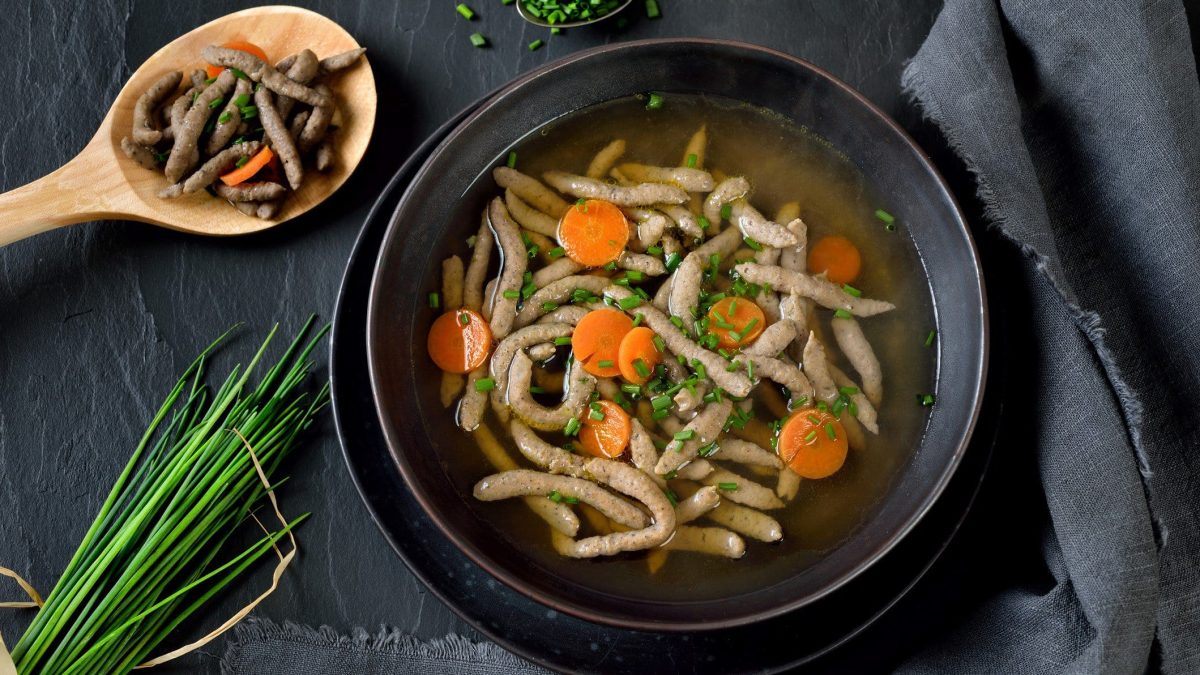 Draufsicht: Auf einem schwarzem Untergrund steht ein dunkler Teller Rindersuppe mit Leberspätzle. Drum herum liegen frischer Schnittlauch un ein Kochlöffel, auf dem Leberspätzle mit Karottenscheiben liegen.
