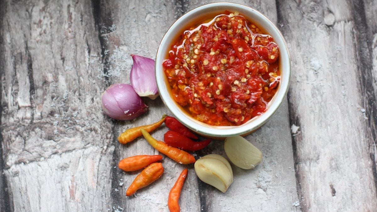Eine weiße Schale Sambal Bawang indonesisches Sambal auf hellem Holz. Im Bild Schalotten, rote Chilischoten und geschälte Knoblauchzehen. Draufsicht.