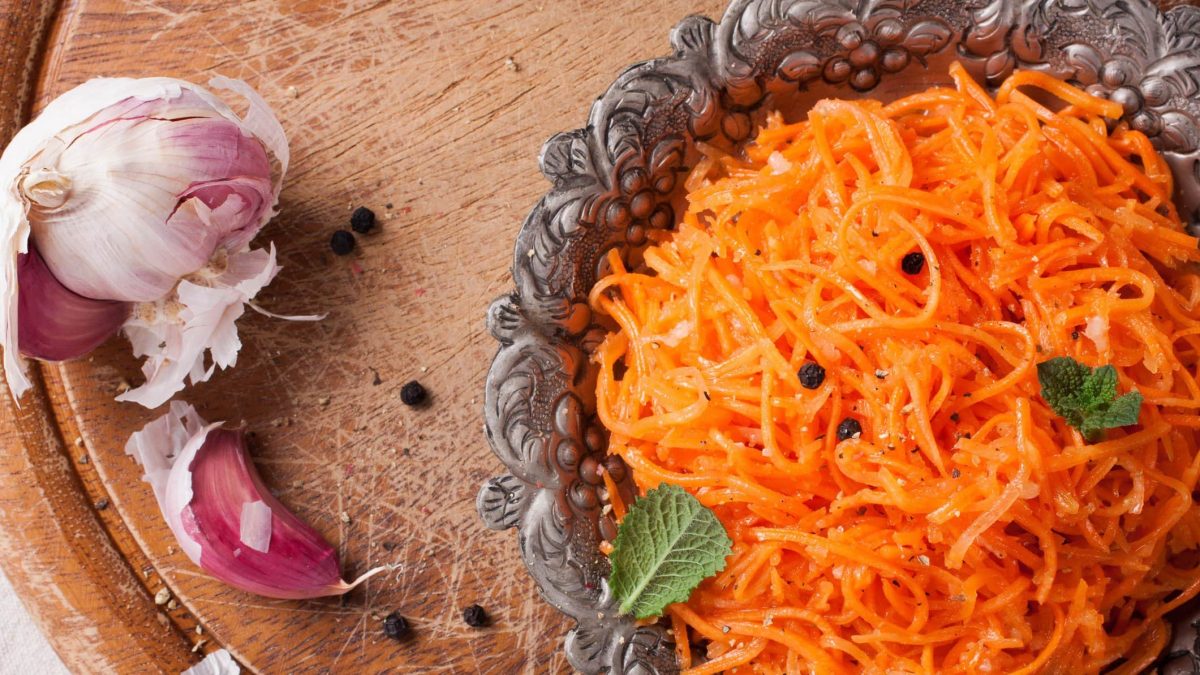Draufsicht: Auf einem runden Holzbrettchen liegt ein orientalisch anmutender Teller mit einer scharfen Scharfe Möhren-Pasta Aglio e Olio. Daneben liegen schwarze Pfefferkörner und frischer Knoblauch.