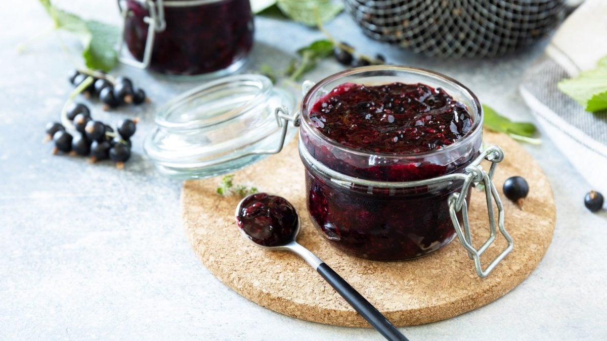 Draufsicht, schräg: Ein Einmachglas Schwarze Johannisbeeren-Marmelade steht auf einem Brettchen auf einem hellen Untergrund. Drum herum liegen schwarze Johannisbeeren und Blätter.