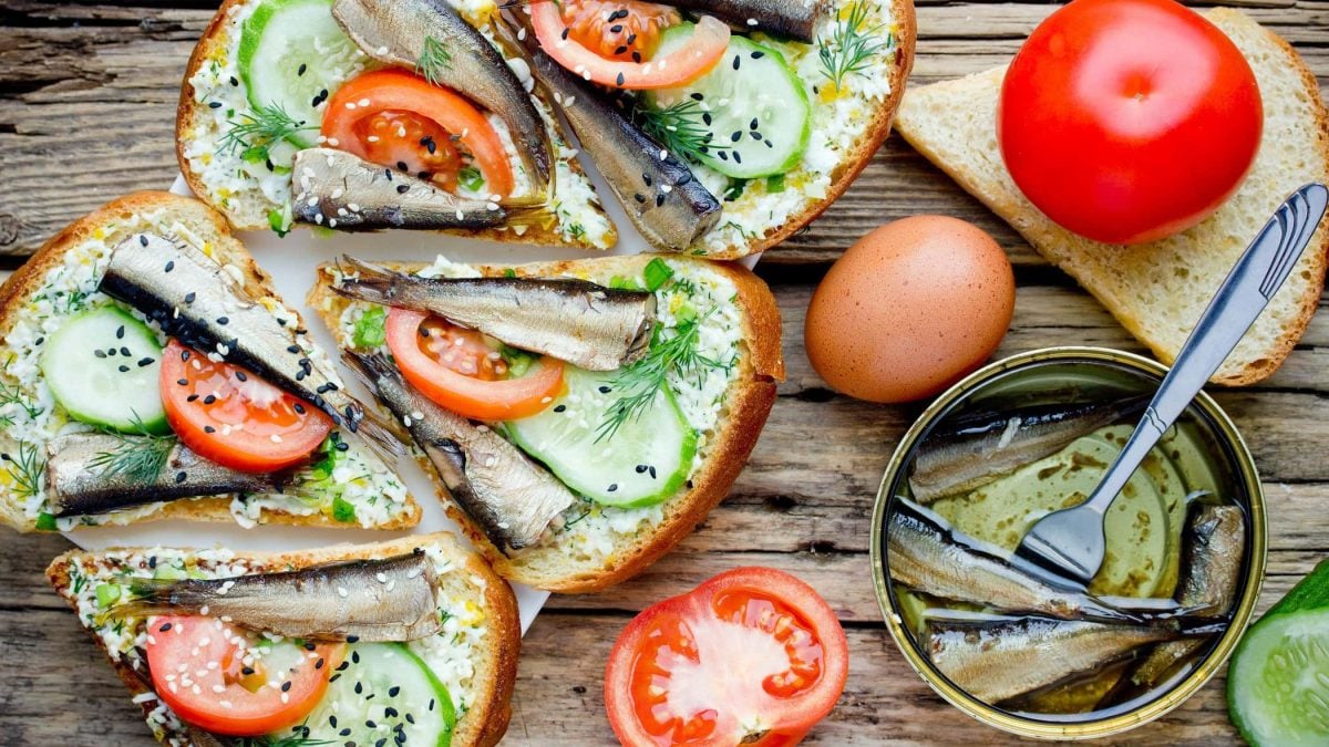 Draufsicht: Sprotten-Sandwich mit Tomaten, Gurken und schwarzem Sesam liegen auf einem Untergrund aus Holz.