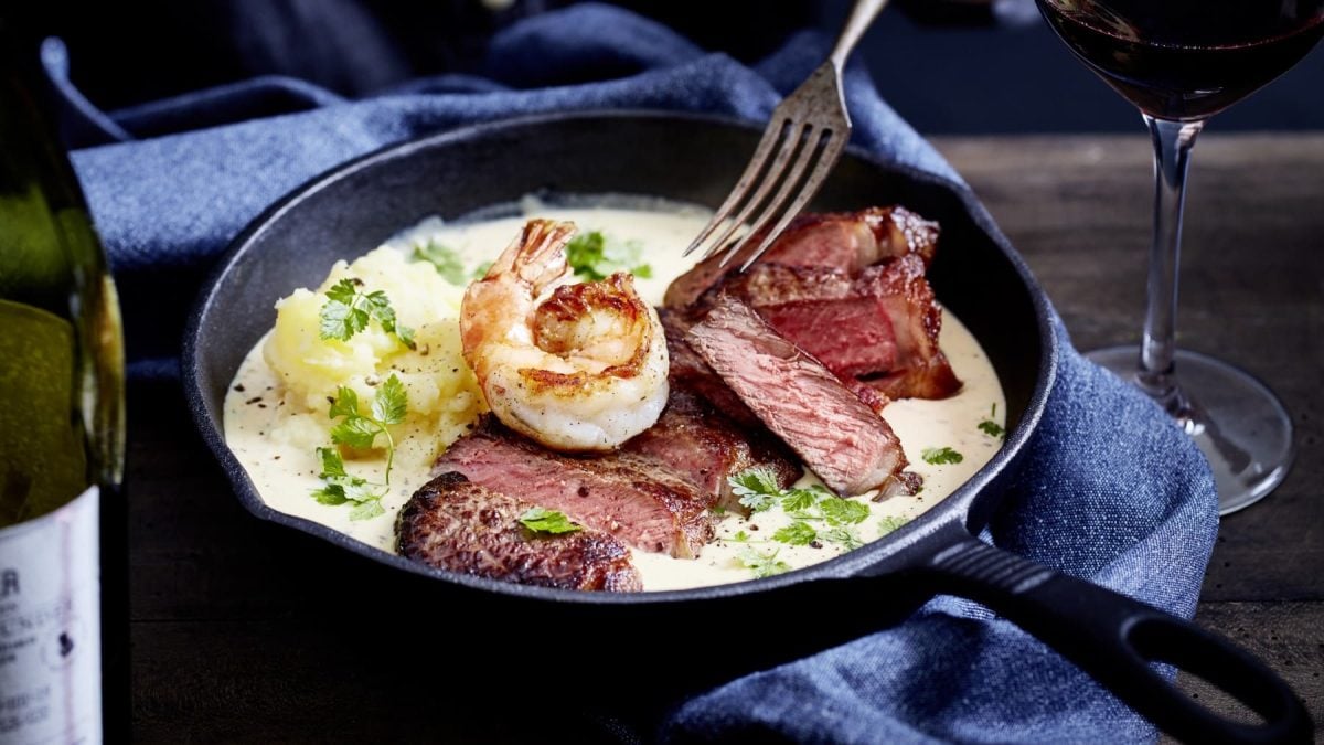 Ein Steak mit Sauce und Garnelen n einer schwarzen Pfanne auf einem dunkelblauen Geschirrtuch. Rechts davon ein Glas Rotwein auf einem hölzernen Hintergrund.