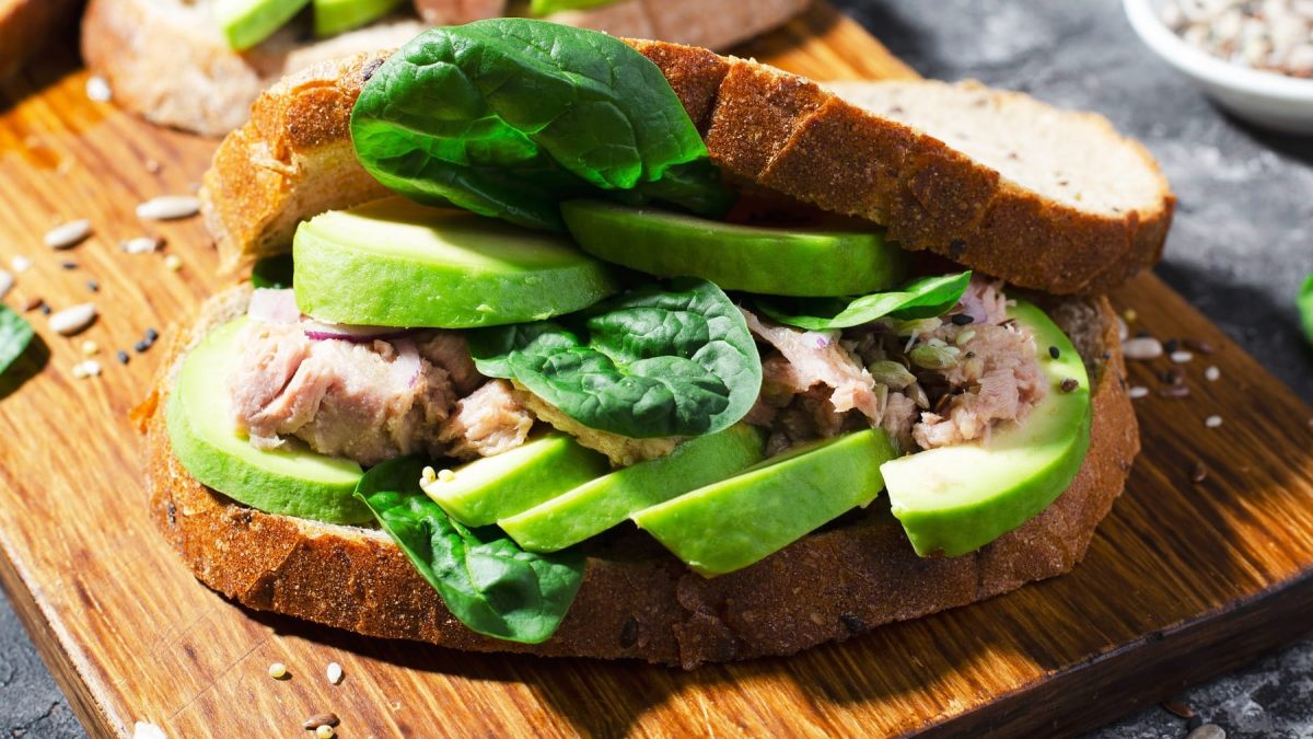 Ein Sandwich mit Avocado, Thunfisch und Babyspinat auf einem Holzbrett und grauem Untergrund.