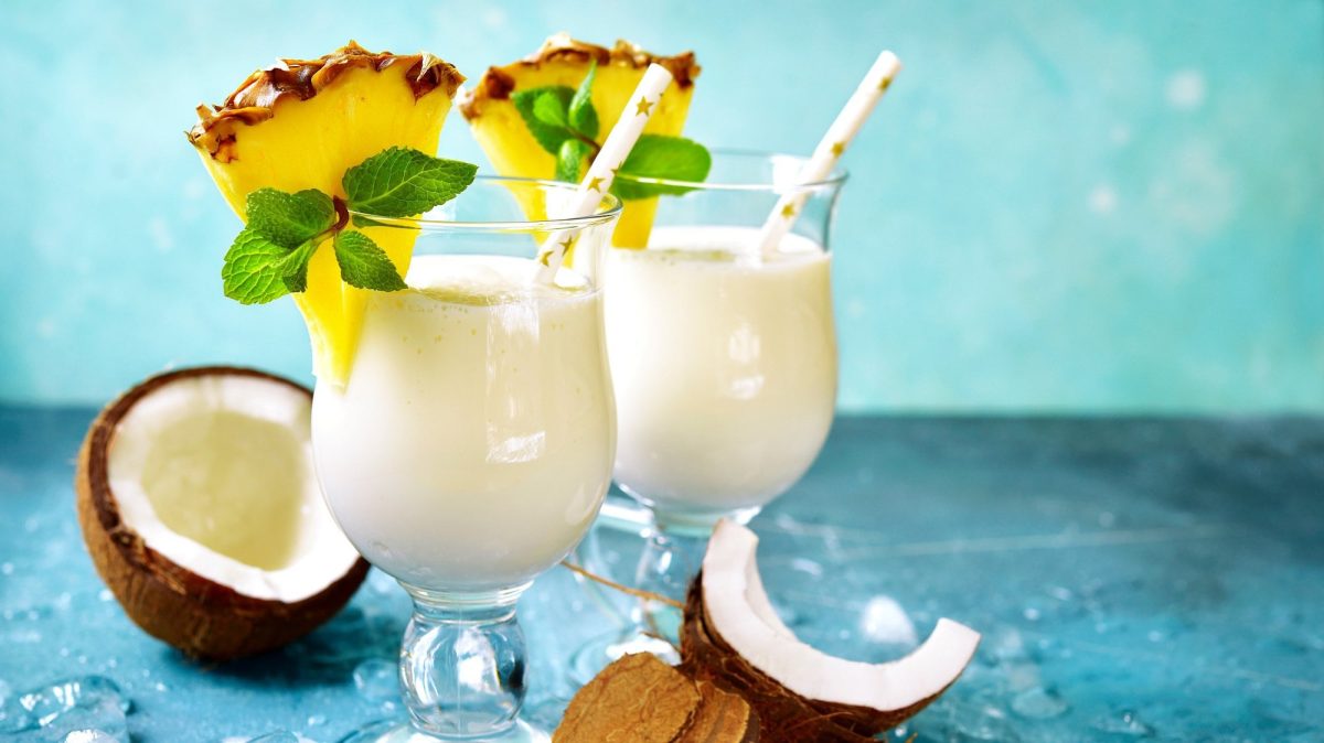 Zwei Gläser vegane Piña Colada, dekoriert mit einer Ananasscheibe und Minze, darin ein Strohhalm, drumherum aufgeschnittene Kokosnüsse.