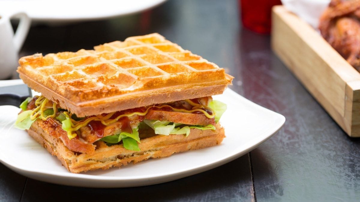 Ein Waffel-Sandwich belegt mit Räuchertofu, Salat, Tomate und Gurke, darauf eine gelbe und rote Sauce, auf einem Teller.