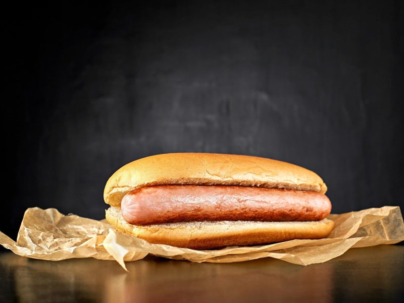 Welche Wurst für Hotdogs? Ein Frankfurter Würstchen im Hotdogbrötchen dem Betrachter zugewandt auf Backpapier vor schwarzem Hintergrund. Frontalansicht.