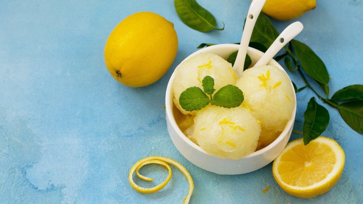Drei Kugeln Zitronensorbet in einer Schale, darin zwei Löffel, drumherum frische Zitronen und MInze.