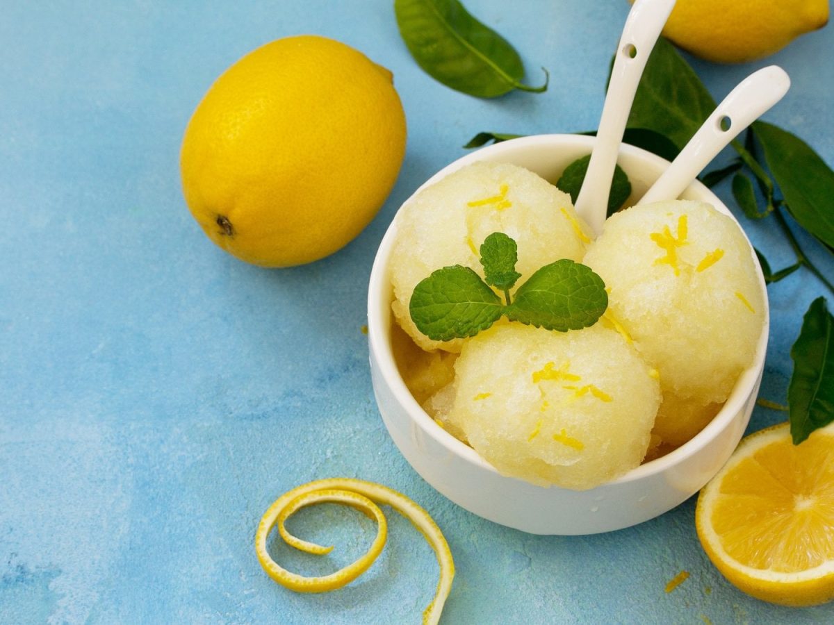 Drei Kugeln Zitronensorbet in einer Schale, darin zwei Löffel, drumherum frische Zitronen und MInze.
