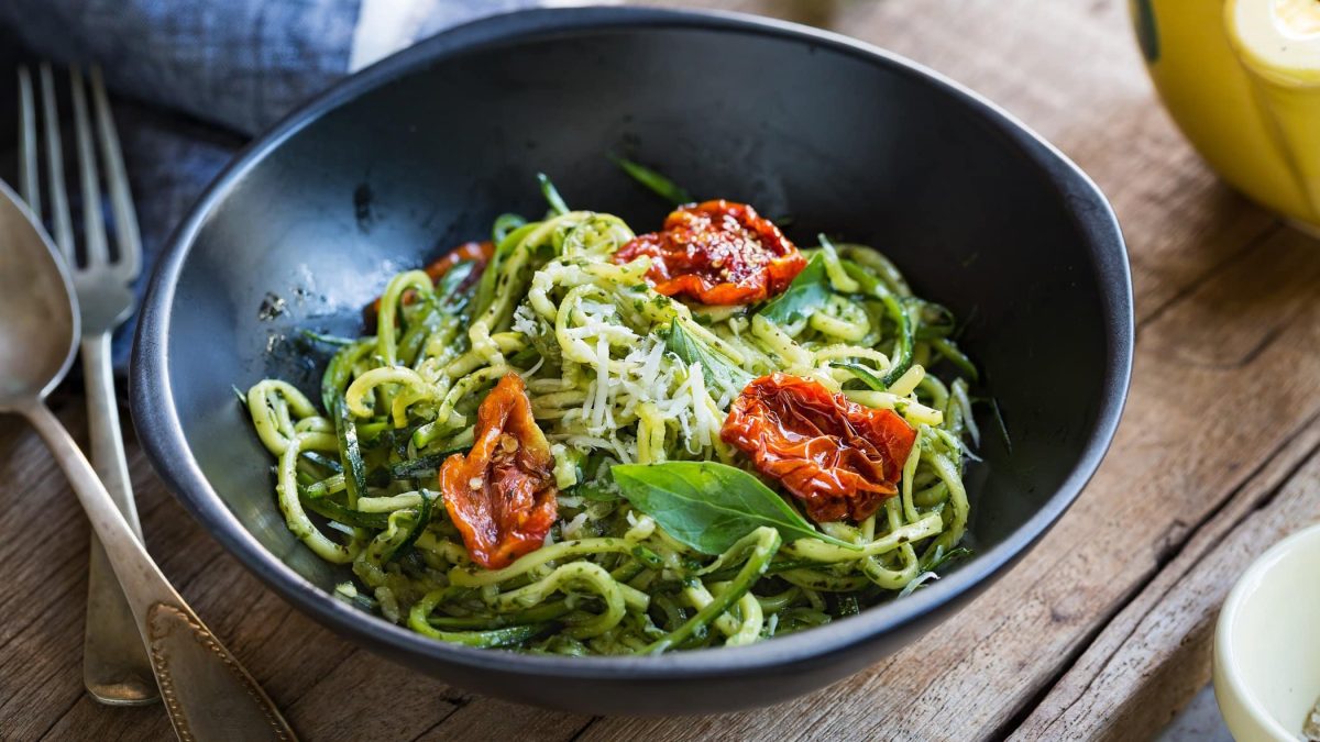 Draufsicht: Eine schwarze Schüssel mit Zucchini-Nudeln mit getrockneten Tomaten steht auf einem Holztisch. Daneben liegt ein Gabel.