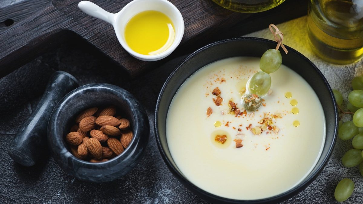 Draufsicht: Dunkler Untergrund, eine runde, dunkle Schale mit einer Ajoblanco. Neben der spanischen Mandelsuppe ist ein Schälchen mit Mandeln sowie ein eine Schüssel mit Olivenöl.