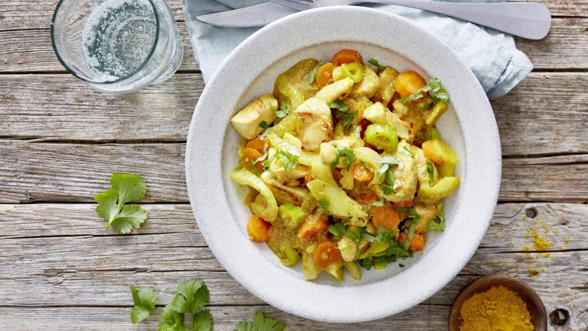 Draufsicht: Amaranth-Fenchel-Pfanne mit Huhn in weißem Teller auf Holzuntergrund, daneben ein Glas und Kräuter verteilt.