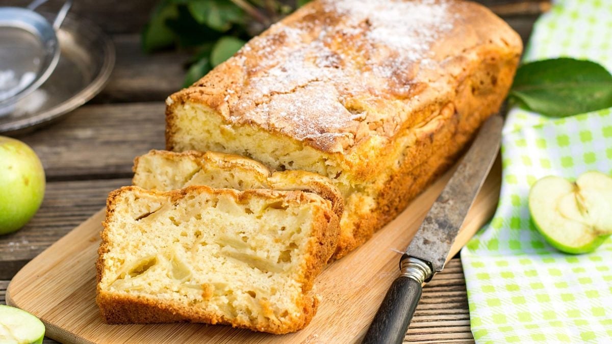 Ein saftiger Apfel-Kastenkuchen auf einem hellen Holzbrett und einem dunklen Tisch mit frischen grünen Äpfeln und einer grün-weiß-karierten Tischdecke.