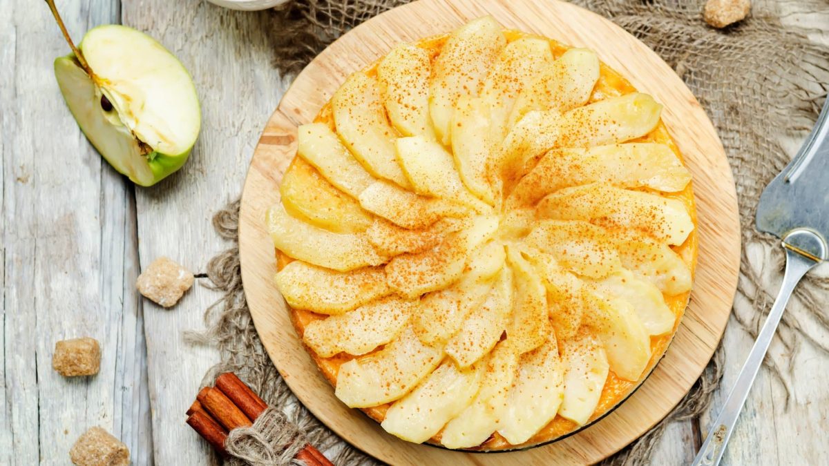 Von-Oben-Ansicht auf gedeckten Apfelmus-Schmandkuchen auf hellem, rustikalem Holz. Daneben liegen Zucker, Apfel und Zimtstangen.