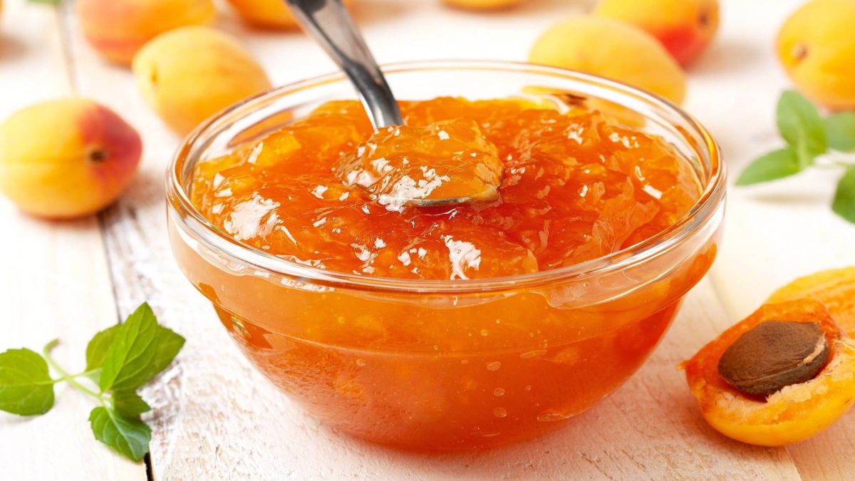 Frontal: Weißer Untergrund, eine Glasschälchen mit Aprikosen-Spritz-Konfitüre sowie frischen Aprikosen.