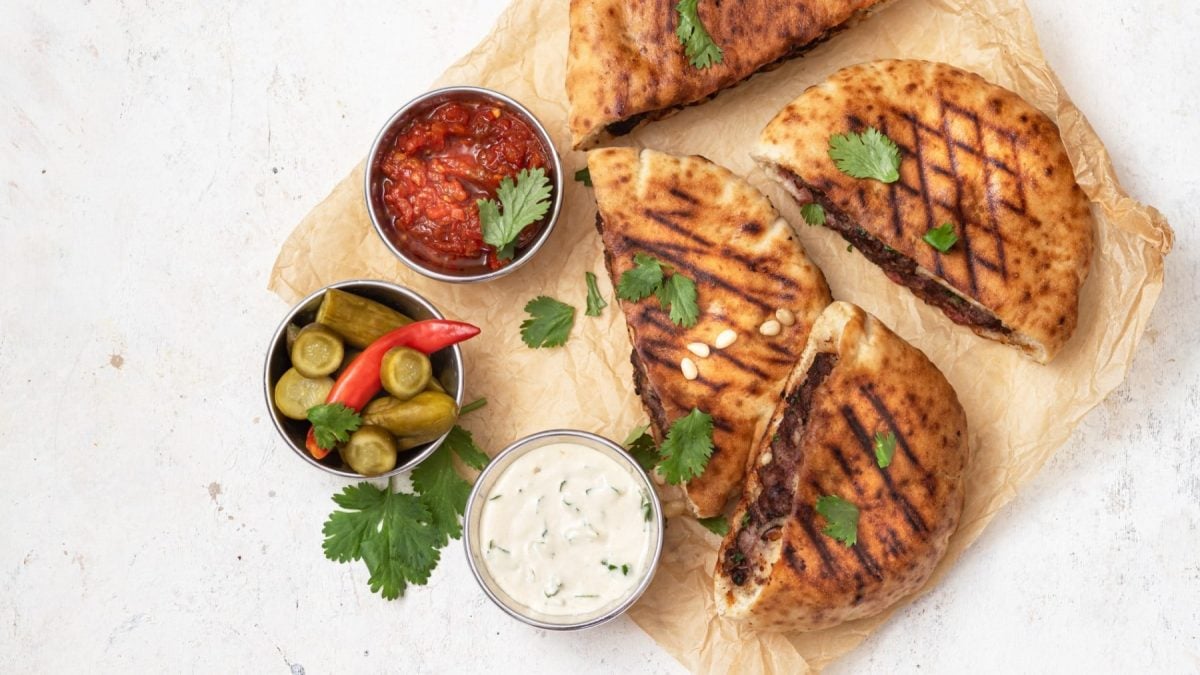 Vegane Arayes auf Backpapier auf einem weißen Hintergrund. Links von den Pita-Broten drei Schälchen gefüllt mit Tahini-Sauce, Salsa und Salzgurken. Alles ist garniert mit frischem Koriander.