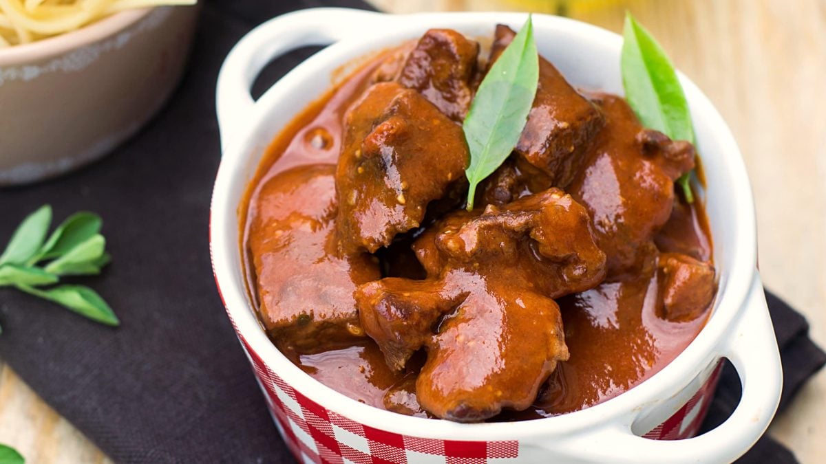 Eine kleine rot-weiß-karierte Suppenschale mit dem Barbecue-Gulasch auf einer schwarzen Tischdecke und einem hellen Holztisch.