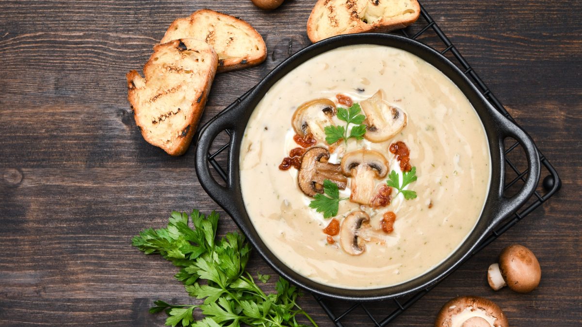 Draufsicht: Dunkler Untergrund, eine dunkle Schüssel bayerische Kümmelrahmsuppe mit Champignons. Daneben liegen frische Petersilie, Brotscheiben und Pilze.