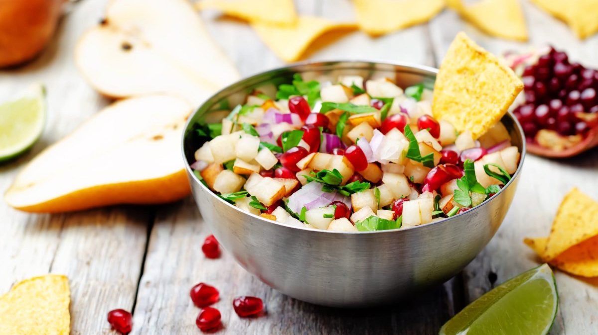 Eine Metallschüssel mit Birnen-Granatapfel-Salsa mit Koriander und roten Zwiebeln, darin steckt ein Nacho-Chip. Drumherum liegen Zutaten wie halbierte Birnen und ein aufgeschnittener Granatapfel.