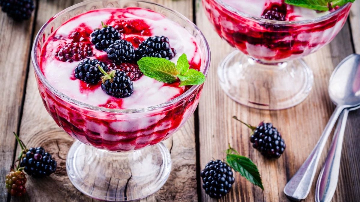 Schräge Draufsicht: Zwei Dessert-Gläser mit einer Brombeer-Sahne-Joghurt Mischung auf einem Holztisch.