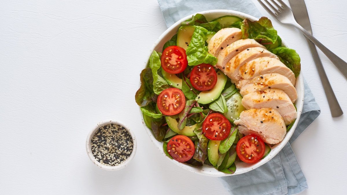 Draufsicht: Auf hellblauem Untergrund steht eine Grüne Bowl mit Putenfilet und Mandel-Dressing. Daneben lieben eine Besteck und ein Schälchen mit Sesamsamen.