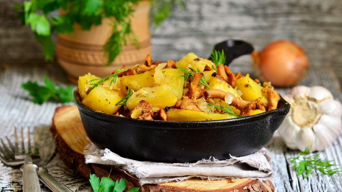 Eine Schüssel Bratkartoffeln mit Pfifferlingen und frischen Kräutern, drumherum liegen Knoblauch, Kräuter und Pilze.