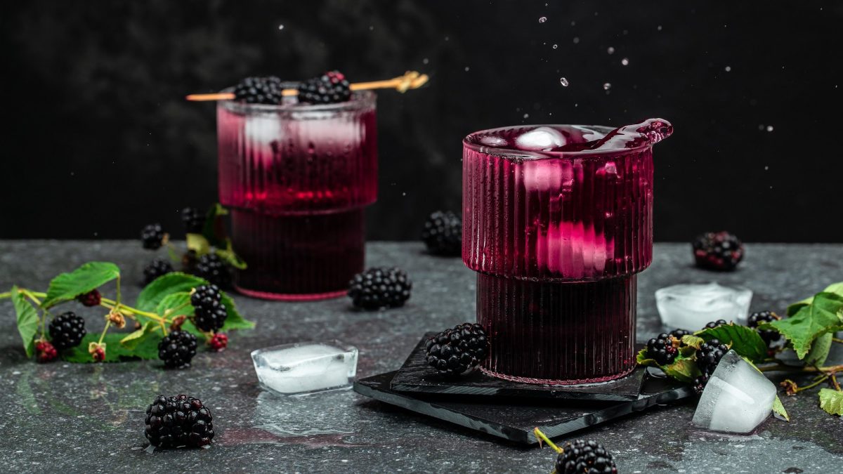 Zwei Gläser Brombeer-Mojito mit Eiswürfeln und Minze, dekoriert mit Brombeeren auf einem Zahnstocher, dunkle Umgebung, drumherum liegen Eiswürfel, Minze und Brombeeren.
