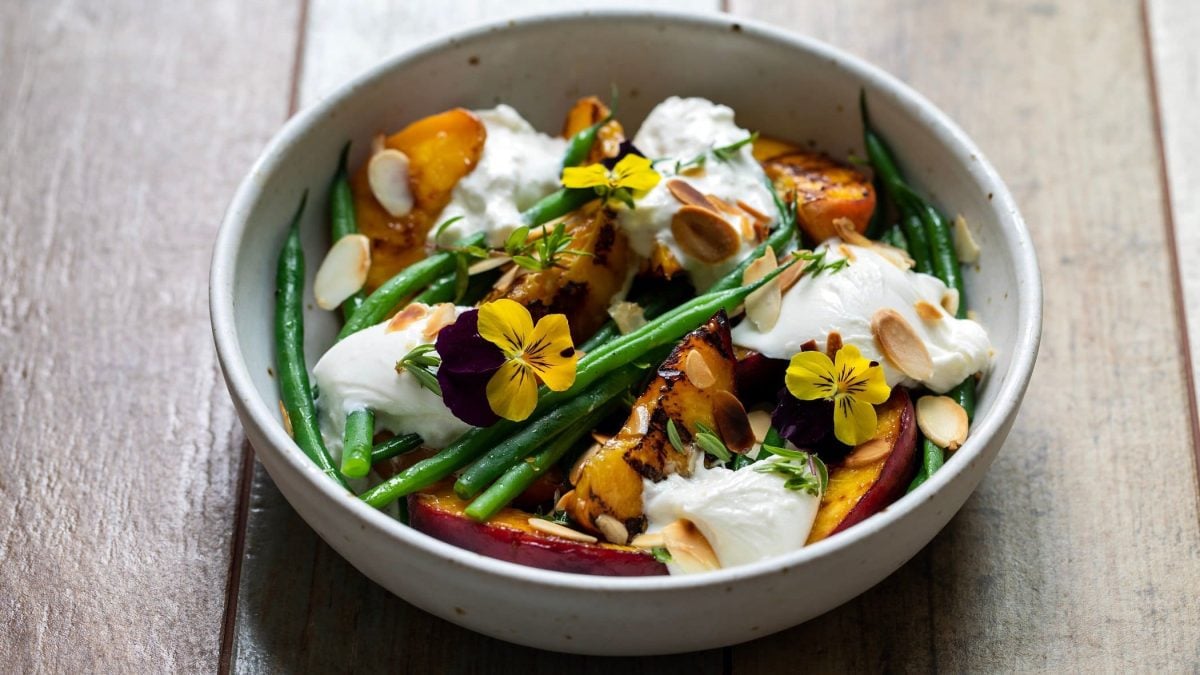Schräge Draufsicht: Eine runde Bowl mit einem Burrata-Salat mit gegrillten Pfirsichen und grünen Bohnen auf einem Untergrund aus Holz.