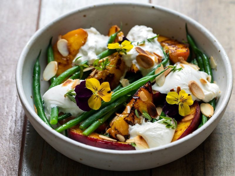 Schräge Draufsicht: Eine runde Bowl mit einem Burrata-Salat mit gegrillten Pfirsichen und grünen Bohnen auf einem Untergrund aus Holz.