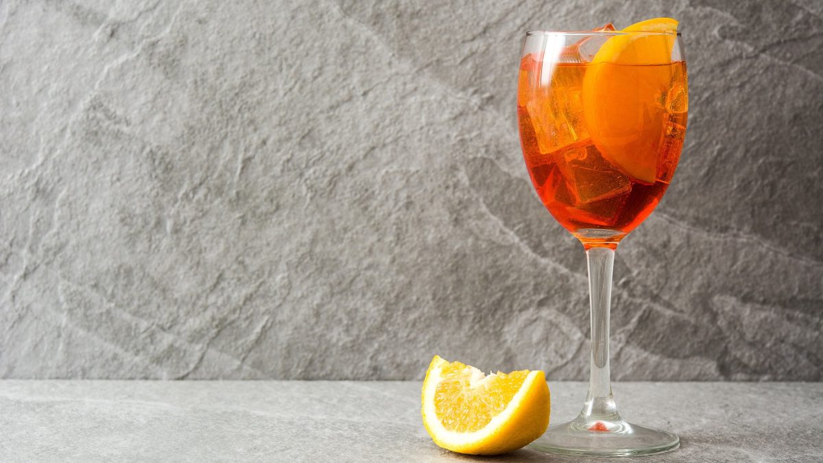 Ein Glas Cappelletti Spritz mit Eis und Zitronenscheiben, daneben liegt eine Zitronenspalte.
