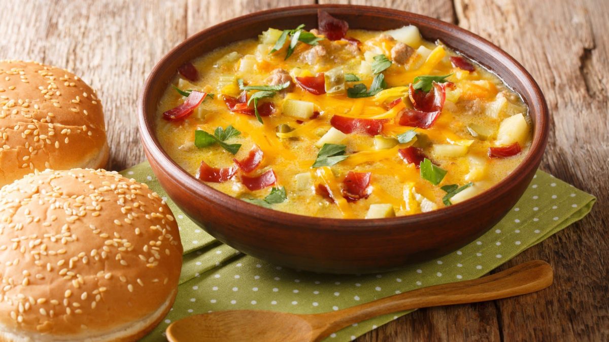 Cheeseburger Suppe mit Hackfleisch, Käse und Kartoffeln auf Serviette und Holztisch. Daneben zwei Burgerbrötchen. Unten im Bild ein Holzlöffel. Draufsicht.