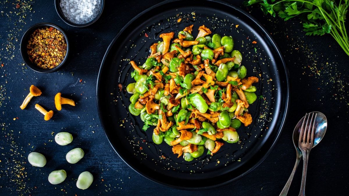 Edamame Salat mit sautierten Pfifferlingen in schwarzem Teller auf dunklem Untergrund. Im Bild rohe Bohnen, Pfifferlinge, Petersilie und, Besteck und Gewürze in schwarzen Schüsseln. Draufsicht