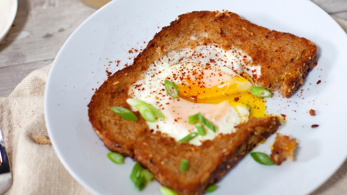 Eine Brotscheibe mit dem Egg in a Hole zusammen mit Chilipulver und Frühlingslauch in sehr naher Aufnahme.