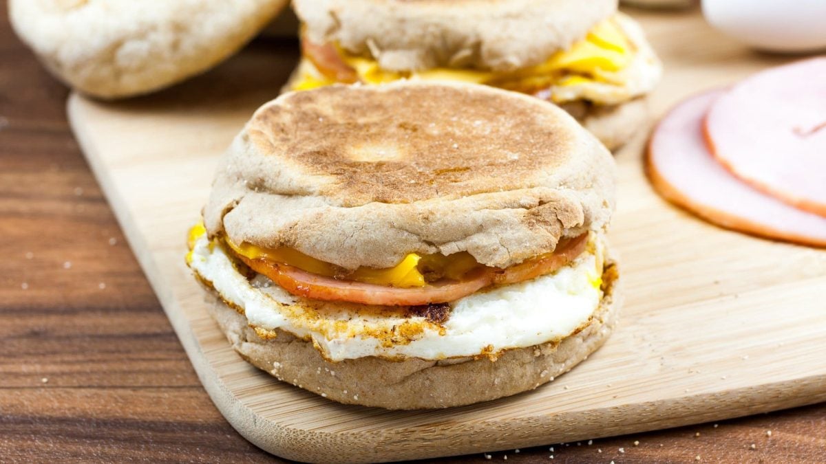 Egg McMuffin mit Bacon auf Holzplatte. Im Hintergrund Kochschinken und weitere englische Muffins auf Holztisch. Draufsicht.