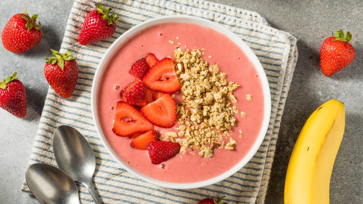 Eine weiße Schüssel mit der Erdbeer-Joghurt-Bowl auf einem grauen Küchentisch mit Erdbeeren und Banane.