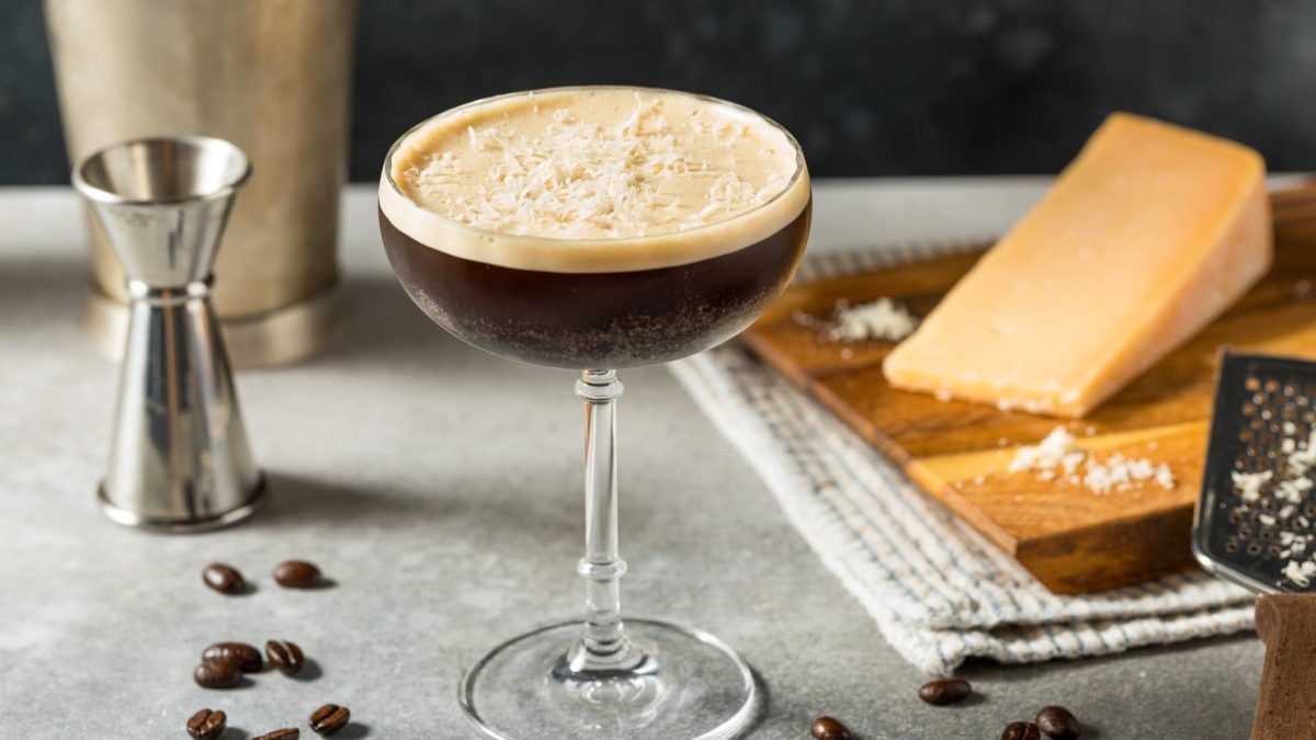 Ein grauer Tisch mit dem Espresso-Martini. Im Hintergrund unscharf Parmesan und ein silberner Jigger zu erkennen.