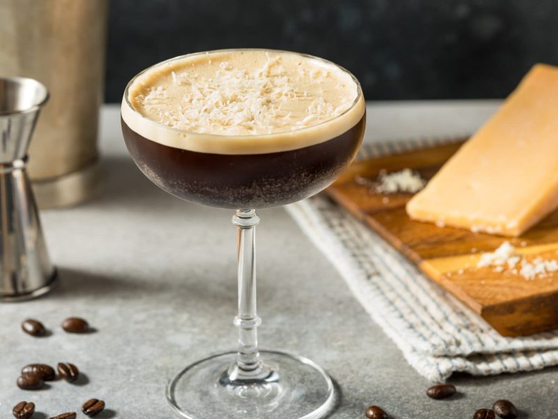 Ein grauer Tisch mit dem Espresso-Martini. Im Hintergrund unscharf Parmesan und ein silberner Jigger zu erkennen.