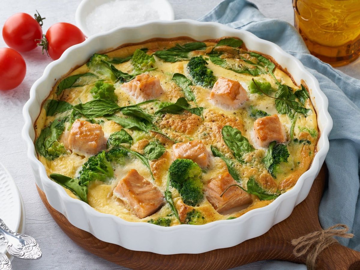 Draufsicht: eine Frittata mit Lachs und Brokkoli in einer weißen Auflaufform auf einem Holzbrett.