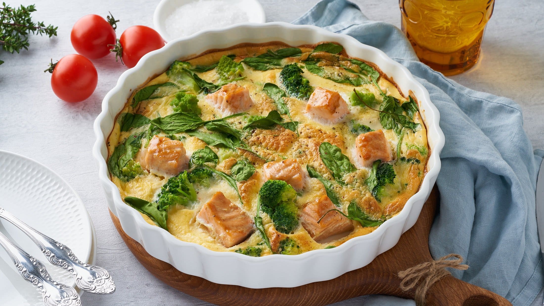 Draufsicht: eine Frittata mit Lachs und Brokkoli in einer weißen Auflaufform auf einem Holzbrett.