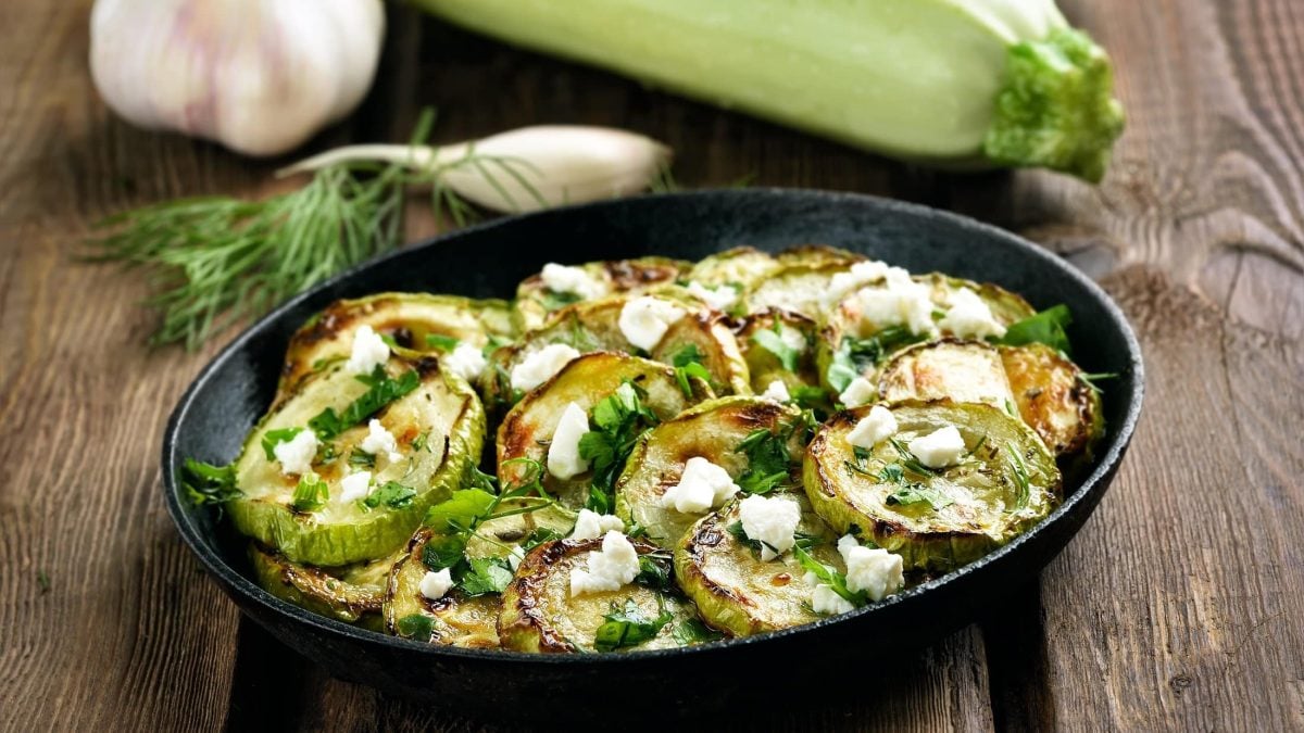 Schräge Draufsicht: Eine Pfanne frittierte Zucchini mit Feta auf einem Untergrund aus Holz.