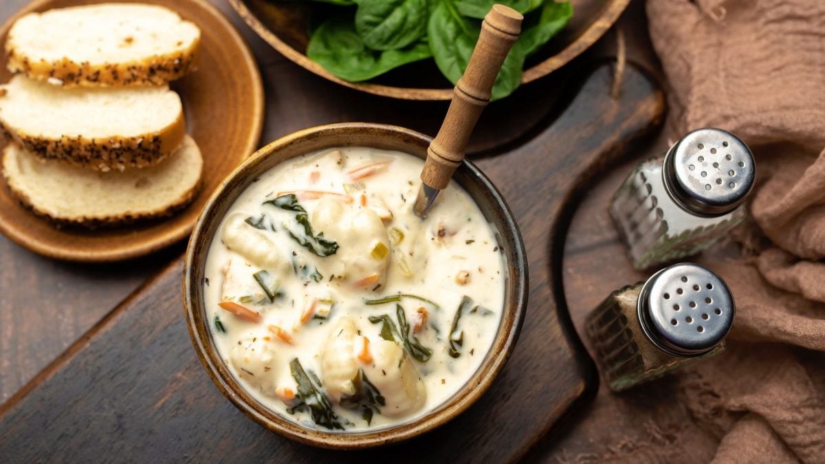 Eine braune Holzschüssel mit der Gnocchi-Suppe auf einem dunklen Holzbrett von oben fotografiert. Daneben Salz- und Pfefferstreuer sowie Brotscheiben und Babyspinat.