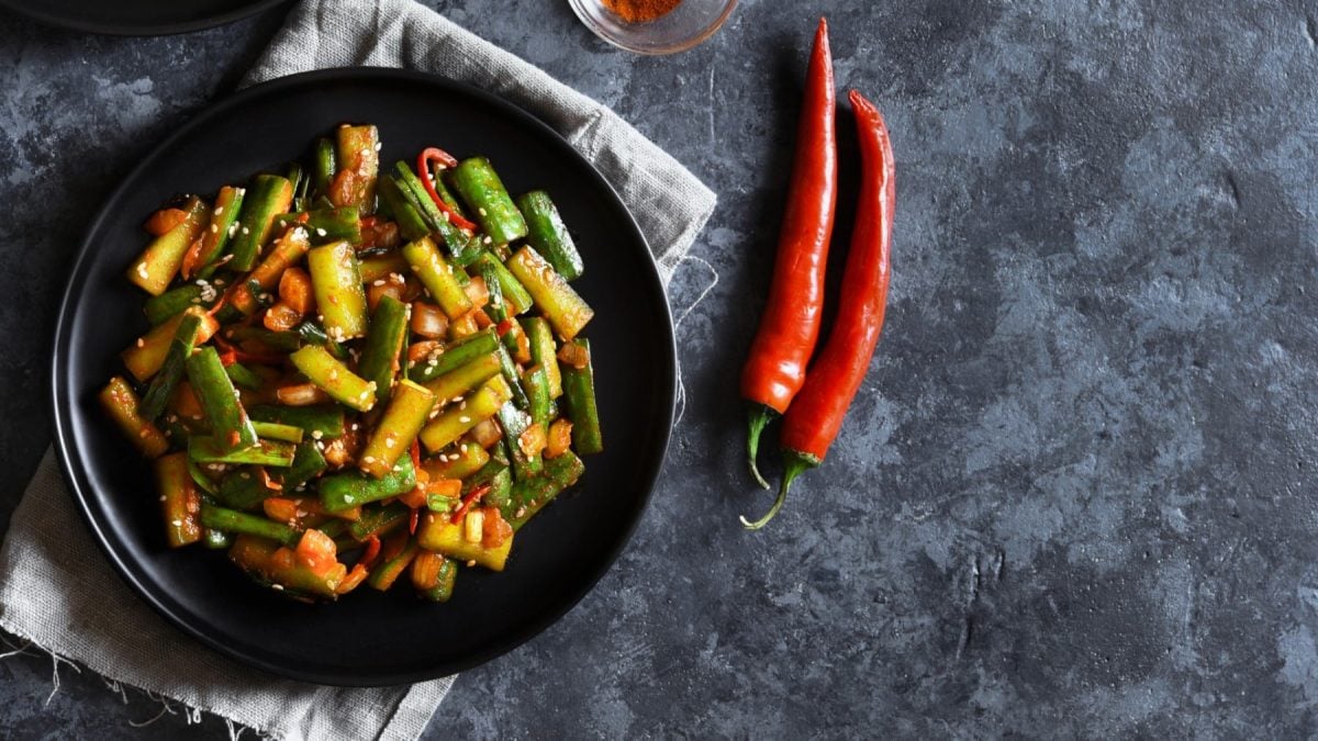 Gurken-Kimchi in einer schwarzen Bowl auf einem grauen Geschirrtuch auf einem dunkelgrauen Hintergrund. Rechts davon zwei rote Chilischoten.